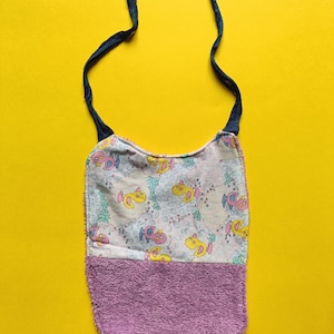Può includere: Un bavaglino per bambini con una parte superiore fantasia e una parte inferiore in spugna viola. La parte superiore presenta un motivo ripetuto di personaggi dei cartoni animati gialli e rosa su uno sfondo chiaro. Lacci blu scuro attaccati in alto.