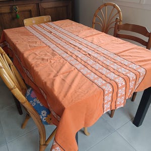 Peut inclure: Une nappe rectangulaire orange ornée d'un panneau central vertical avec une broderie géométrique blanche et noire complexe. Le tissu présente une texture lisse et retombe sur les bords d'une table, révélant un motif répétitif.