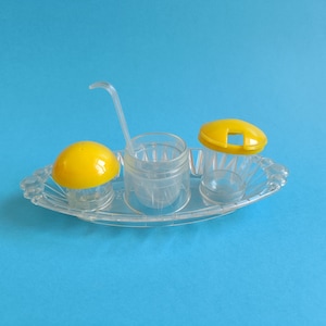 Peut inclure: Un ensemble de condiments en plastique transparent avec des couvercles jaunes et une cuillère de service. L'ensemble comprend un salière, un poivrier et un récipient pour le sucre ou d'autres condiments. L'ensemble repose sur un plateau transparent de forme ovale.