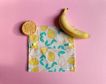 May include: A reusable sandwich bag with a lemon pattern, a banana, and a cracker on a pink background. The bag is beige with yellow lemons and teal leaves. The banana is yellow and the cracker is round and golden.