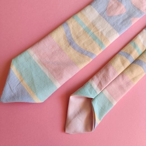 Peut inclure: Une cravate aux couleurs pastel avec un motif à rayures et à cœurs. La cravate présente des teintes bleu clair, rose, jaune et vert. Le fond est de couleur rose uni.