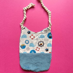 Può includere: Un bavaglino per bambini a tema nautico, con balene, barche e salvagenti su sfondo bianco. La metà inferiore è in morbida spugna blu e il cinturino è a righe. Il bavaglino è per neonati.