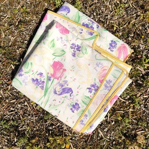 Peut inclure: Deux serviettes en tissu à motifs floraux sur fond blanc, avec des fleurs roses, violettes et vertes. Les serviettes ont une bordure jaune. Les serviettes sont pliées et empilées l'une sur l'autre.