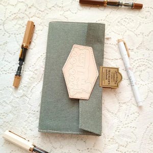 May include: A sage green notebook cover with a light tan leather patch featuring a vehicle design. Several pens in shades of brown and white are scattered around the notebook. A small, rectangular tag with text is also present.