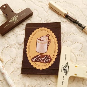 May include: A brown notebook cover with a latte design. The cover features a cream-colored oval with a drawing of a latte and the word "Latte" below it. Two brown and white binding clips and a pen are also visible.