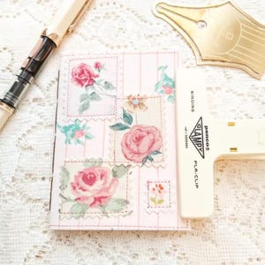 May include: A pink floral journal with rose illustrations, a gold pen, and a white binding clip. The journal features a repeating pattern of roses in various sizes and colors, with a stitched border.