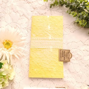 May include: A rectangular yellow notebook cover with a textured floral pattern and a horizontal white lace trim across the center. A small brown rectangular tag is attached to the side, featuring the text TRAVELER'S COMPANY JAPAN TRAVELER'S notebook.