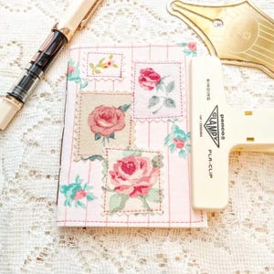 May include: A floral patterned notebook with pink and green rose designs. A white and black fountain pen and a white binder clip are next to the notebook. The binder clip has the text "BINDING CLAMPY PLA-CLIP".