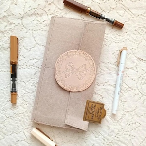 May include: A beige notebook with a round, embossed bow design. The notebook is surrounded by brown and white pens, and a small tag that reads "TRAVELER'S COMPANY" is also present. The items are arranged on a white lace surface.