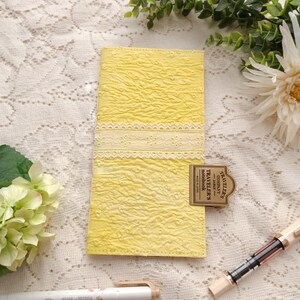May include: A rectangular yellow notebook cover featuring a textured fabric surface with a horizontal white eyelet lace trim across the center. A brass-colored metal clip is attached to the side, embossed with the text TRAVELER'S COMPANY JAPAN.