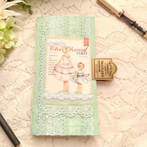 May include: A light green fabric notebook cover decorated with white lace trim and a vintage-style paper insert featuring two illustrated figures in dresses. A brass-colored metal clip is attached to the side with the text Traveler's Company Japan.
