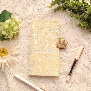 May include: A cream-colored lace notebook cover featuring a floral pattern and a brass-toned metal clip embossed with the text TRAVELER'S COMPANY JAPAN TRAVELER'S notebook MADE IN JAPAN, accompanied by a white pen and a clear-barreled fountain pen.