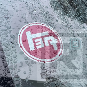 May include: A red circle sticker with a white "TR" logo on a car windshield covered in raindrops.