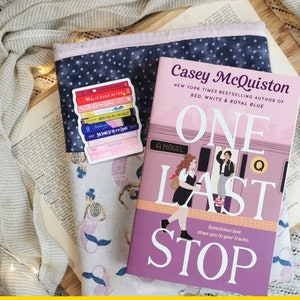 以下が含まれることがあります： 「One Last Stop」というタイトルのピンク色のペーパーバック。ニューヨーク・タイムズでベストセラー作家であるケイシー・マクィストン著。ケイシー・マクィストンは「Red, White & Royal Blue」の著者としても知られています。表紙には、地下鉄の電車に乗っている2人の女性のイラストが描かれています。本は、人魚のデザインのブックカバーと、本の積み重ねのステッカーが貼られたテーブルの上に置かれています。