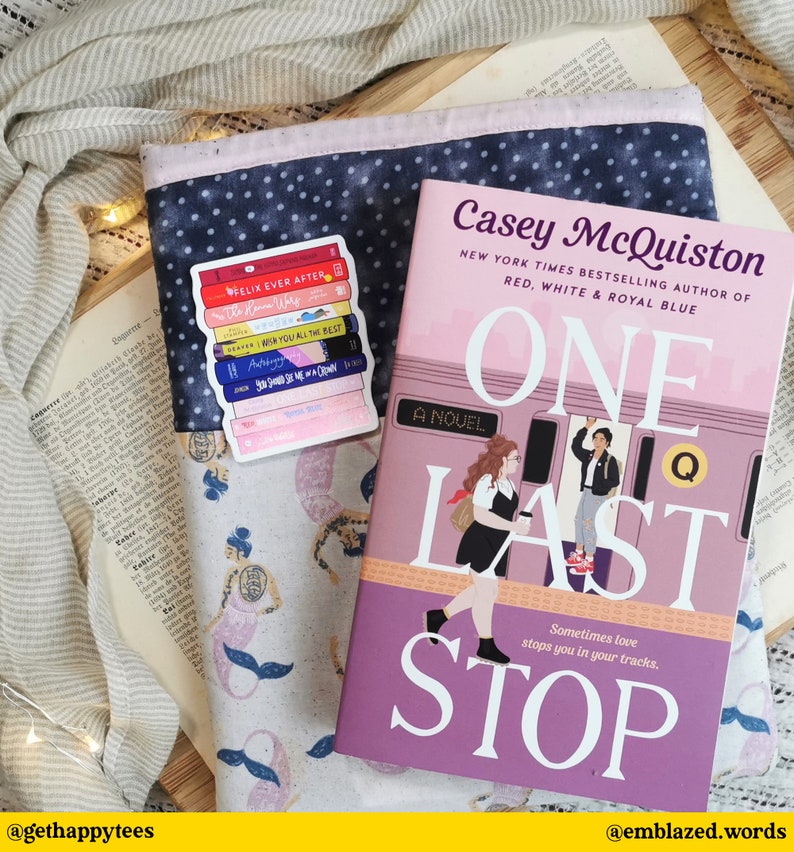 以下が含まれることがあります： 「One Last Stop」というタイトルのピンク色のペーパーバック。ニューヨーク・タイムズでベストセラー作家であるケイシー・マクィストン著。ケイシー・マクィストンは「Red, White & Royal Blue」の著者としても知られています。表紙には、地下鉄の電車に乗っている2人の女性のイラストが描かれています。本は、人魚のデザインのブックカバーと、本の積み重ねのステッカーが貼られたテーブルの上に置かれています。