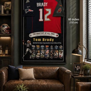 May include: Framed artwork featuring a split football jersey, one side navy blue, the other red, with "BRADY 12" prominently displayed. The artwork is 36 inches (91 cm) wide and 60 inches (152 cm) tall, with text that reads "THE GREATEST OF ALL TIME".