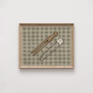 May include: A framed print with a brown and white gingham pattern background. A wooden clothespin and a safety pin are placed on the gingham pattern.