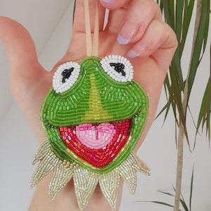 May include: A beaded Kermit the Frog decoration. The ornament has a green face with white and black eyes, a pink and red mouth, and a gold beaded collar. A beige ribbon is attached for hanging.