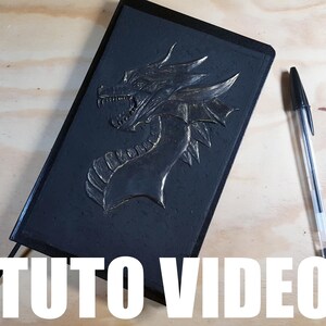 Może przedstawiać: Czarny notatnik z reliefem smoka i czarnym długopisem. Smok jest szczegółowy ze złotymi akcentami. Tekst "TUTO VIDEO" znajduje się na dole obrazu.