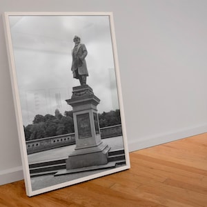 May include: Black and white framed print of a statue of a man in a long coat, standing atop a detailed pedestal. The artwork is leaning against a white wall and a wooden floor. The background shows trees and a cloudy sky.