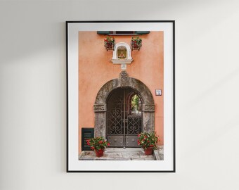 Peut inclure: Impression encadrée d'une porte arquée avec des grilles en fer forgé. Le bâtiment couleur pêche présente une arche en pierre, un petit sanctuaire religieux et des fleurs rouges en pot. Un cadre noir entoure l'image.