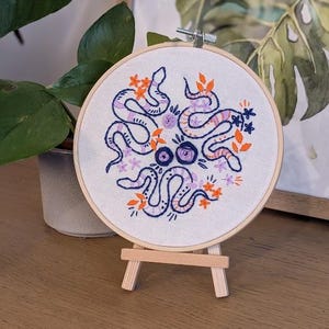May include: A round embroidery hoop featuring a purple and blue snake design with orange and purple floral accents. The embroidery is displayed on a small wooden easel, with a plant and framed artwork in the background.
