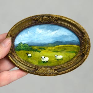 Puede incluir: Miniatura ovalada en un marco dorado ornamentado. La obra representa una escena pastoral con tres ovejas pastando en un campo verde bajo un cielo azul con nubes. El marco tiene detalles de filigrana.