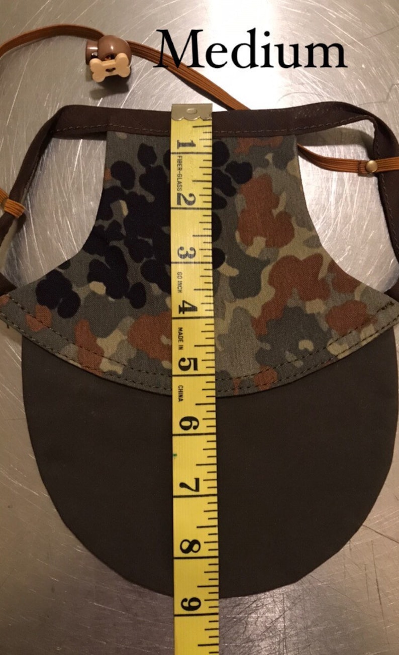 May include: A brown and green camouflage dog sun hat with a brown brim. The hat is measured with a yellow tape measure, showing a size of 6 inches.