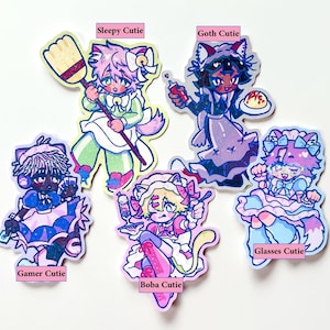 Peut inclure: Six autocollants colorés représentant des personnages de style anime avec des oreilles de chat. Chaque autocollant a un design unique et les textes : "Sleepy Cutie", "Goth Cutie", "Gamer Cutie", "Boba Cutie", et "Glasses Cutie".