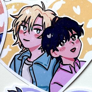 May include: A heart-shaped sticker with two anime characters. The character on the left has blonde hair and is wearing a blue jacket. The character on the right has black hair and is wearing a pink shirt. The background is yellow with white hearts and stars.