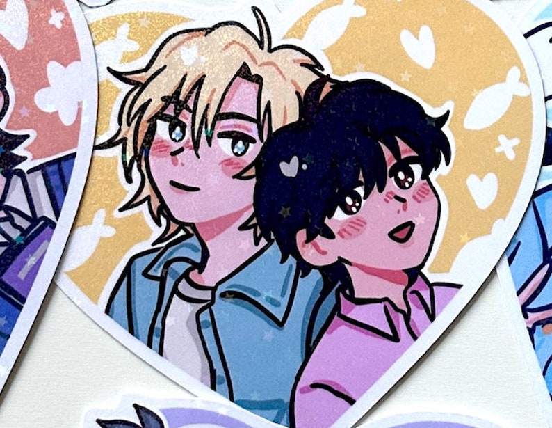 May include: A heart-shaped sticker with two anime characters. The character on the left has blonde hair and is wearing a blue jacket. The character on the right has black hair and is wearing a pink shirt. The background is yellow with white hearts and stars.