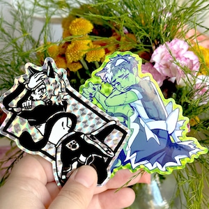 May include: Two holographic stickers featuring cartoon characters. One sticker is black and white and features a creature with a hat and a tail. The other sticker is green and blue and features a creature with a hat and a tail.