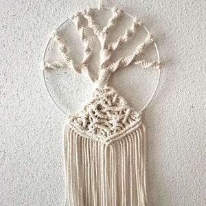 Peut inclure: Suspension murale en macramé crème représentant un arbre. L'arbre est réalisé avec de la corde nouée, inséré dans un cercle en métal blanc. La conception comprend une base texturée détaillée et de longues franges.