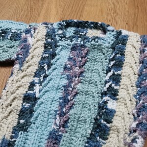 Peut inclure: Un pull en crochet pour bébé avec un motif rayé dans les tons de bleu, vert, blanc et beige. Le pull a une fermeture boutonnée au niveau du cou.
