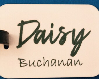 May include: A luggage tag with the name "Daisy Buchanan" printed in dark green script. The tag is rectangular with rounded corners and a black leather strap. The background is a solid blue.