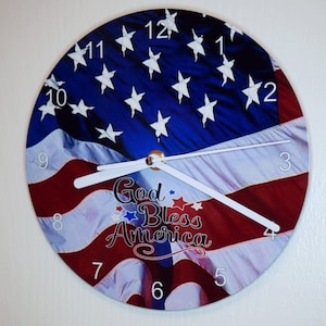 May include: A round clock with a white face and black numbers. The clock face features a waving American flag with the words "God Bless America" in the center.