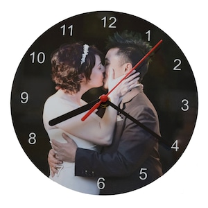 May include: Round wall clock featuring a romantic image of a couple kissing. The clock face displays a photograph of the couple, with black hands and red second hand. White numerals mark the hours against a black background.