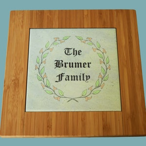 May include: A light brown bamboo trivet with a square ceramic tile insert. The tile has a light blue background with a green wreath and the text "The Brumer Family" in black.