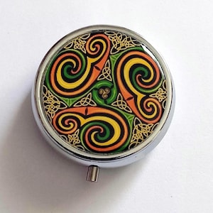 May include: A circular, silver-coloured pill box with a vibrant Celtic knot design. The design incorporates swirling orange, yellow, and green patterns with gold detailing. The pill box has a small, rectangular clasp.