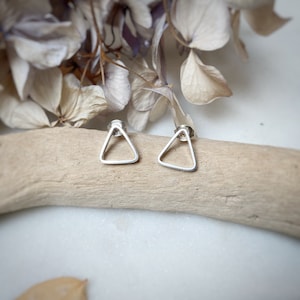 Puede incluir: Un par de pendientes de plata en forma de triángulo con un centro hueco. Los pendientes se muestran sobre una superficie de madera marrón claro.