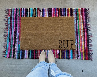May include: A brown coir doormat with the word "SUP" in black lettering. The doormat is surrounded by a colorful, striped rug.