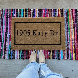 May include: A brown coir doormat with a black border and the text "1905 Katy Dr." printed on it. The doormat is on a colorful striped rug.
