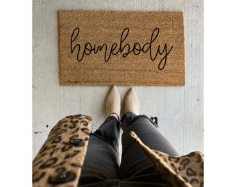 May include: A brown coir doormat with the text "homebody" written in black cursive script.