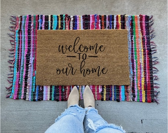 May include: A rectangular coir doormat with the words "welcome to our home" in black script sits on a colorful rag rug. The rug has a fringed edge and a mix of pink, blue, green, and purple stripes.