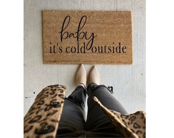 May include: A rectangular doormat made of natural coir fiber. The mat features the phrase "baby it's cold outside" in a black, elegant script. The mat is placed on a concrete surface.