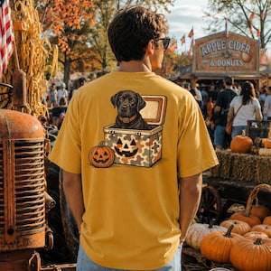 May include: A mustard yellow t-shirt featuring a graphic on the back of a black Labrador retriever sitting inside a camouflage cooler with a jack-o'-lantern face. A separate orange pumpkin sits next to the cooler on the ground.