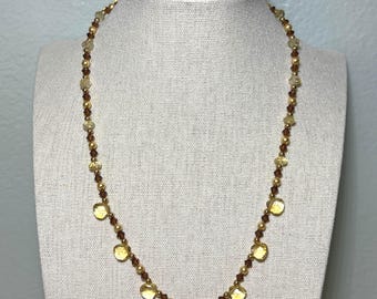 May include: A gold-toned necklace with a variety of beads and pendants. The necklace features small gold beads, amber-colored faceted beads, and larger, flat, yellow pendants. The necklace is displayed on a mannequin.