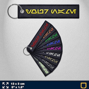 Könnte beinhalten: Ein Set mit 10 schwarzen Schlüsselanhängern mit dem Text "VODZ AKM" in verschiedenen Farben, darunter Rot, Orange, Gelb, Grün, Blau, Lila, Pink und Weiß. Die Schlüsselanhänger sind 13 cm lang und 3 cm breit.