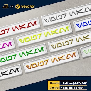 Könnte beinhalten: Ein Set mit sieben Bügel- oder Näh-Patches mit dem Text "VOL07 SKM" in verschiedenen Farben, darunter Rot, Gelb, Grün, Orange und Weiß. Die Patches sind in zwei Größen erhältlich: klein (12 cm x 2 cm) und groß (16 cm x 5 cm).