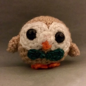May include: A small crocheted owl with large black eyes, a white face, and a teal bow tie. The owl is brown and has orange feet.